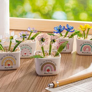 May include: Five small white square ceramic planters, each containing glass flowers and pebbles. The planters feature a rainbow graphic, a red apple icon, and the text "Thank you for helping me grow" with a name printed below the design.
