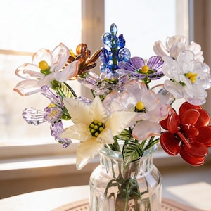 May include: A glass vase holding a vibrant bouquet of handmade flowers. The flowers are in shades of white, red, purple, and blue, with yellow centres. The clear vase displays the flowers, arranged in a natural, artistic manner, in front of a window.