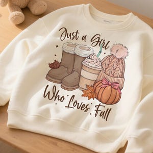 May include: Cream-colored sweatshirt with a fall-themed graphic. The graphic includes brown boots, a coffee drink, a knit hat, and a pumpkin. The text reads "Just a Girl Who Loves Fall."