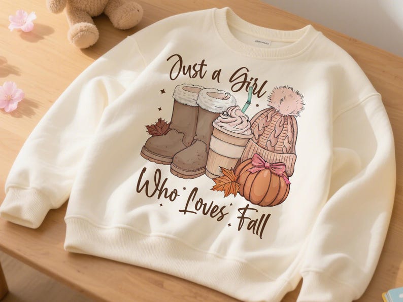 May include: Cream-colored sweatshirt with a fall-themed graphic. The graphic includes brown boots, a coffee drink, a knit hat, and a pumpkin. The text reads "Just a Girl Who Loves Fall."