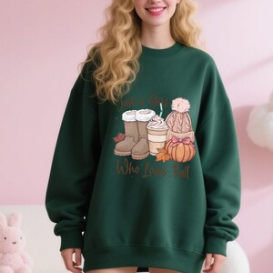 May include: Dark green sweatshirt with a fall-themed graphic. The graphic includes boots, a coffee drink, a hat, and a pumpkin. The text on the sweatshirt reads "Just a Girl Who Loves Fall."