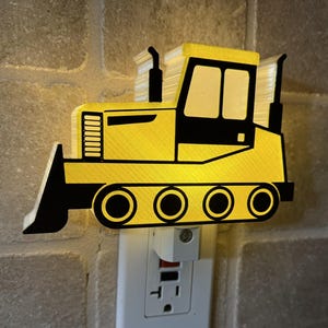 May include: A yellow and black bulldozer night light plugged into a wall outlet. The night light is shaped like a bulldozer with a blade, tracks, and a cab. The light is on, illuminating the bulldozer's design.