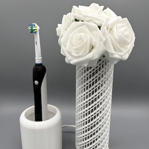 May include: A white electric toothbrush in a white holder next to a white vase with white roses. The vase has a lattice design. The toothbrush is black and white with a blue and green brush head.