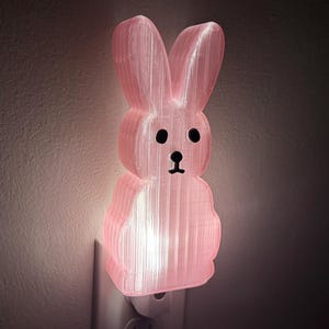 May include: A pink bunny night light plugged into a wall outlet. The bunny has black eyes and a small black nose. The light glows from within the bunny's body, creating a soft, ambient light.