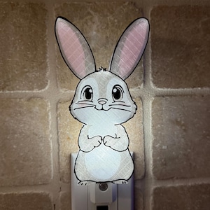 May include: A night light featuring a cartoon bunny with long ears and a sweet expression. The bunny is primarily gray with pink inner ears and is plugged into a wall outlet. The background is a textured, light brown surface.