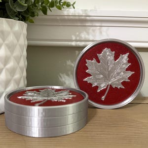 May include: Two round coasters with a silver maple leaf design on a red background. The coasters have a silver rim and are displayed on a wooden surface. A white geometric vase and greenery are in the background.