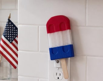 May include: A red, white, and blue popsicle-shaped night light is plugged into a white electrical socket. The kitchen scene includes a framed sign that reads "Land of the *free*" and a small American flag.