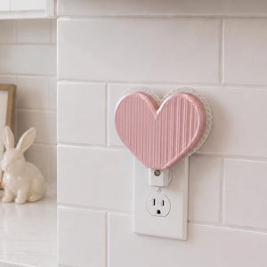 May include: A pink heart-shaped wall plug-in warmer with a vertical ribbed texture and a white lace-like trim edge, plugged into a white electrical outlet on a white tiled wall. The heart features a glossy finish and a rounded, symmetrical shape.