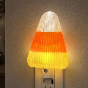 May include: A plug-in night light shaped like a piece of candy corn with a ribbed, translucent texture. The light features horizontal bands of white at the top, orange in the middle, and yellow at the base, emitting a warm, soft glow from the wall.