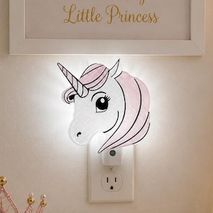 May include: A white and pink unicorn night light plugged into a wall outlet. The unicorn has a black outline and a pink mane and horn. The night light is next to a framed print that says "Dream Big Little Princess".