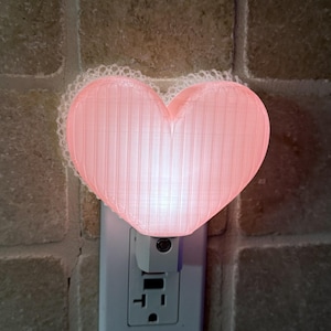 May include: A pink heart-shaped night light with a lace trim, plugged into a wall outlet. The light emits a soft glow, creating a warm ambiance. The night light is a decorative and functional home decor item.