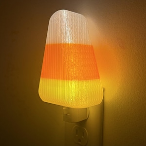May include: A candy corn-shaped night light plugged into a wall outlet. The light is white, orange, and yellow, mimicking the colors of the Halloween treat. The light is on, illuminating the surrounding wall.