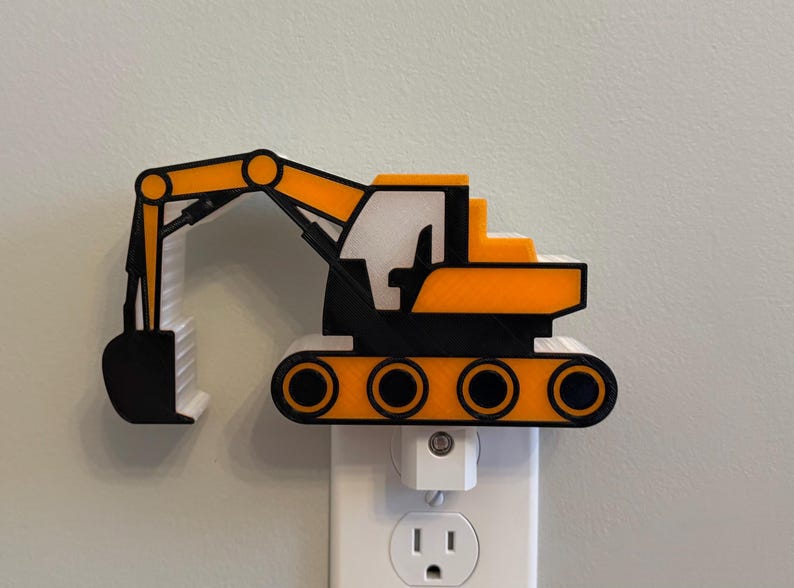 May include: A yellow, black, and white excavator-shaped night light plugged into a standard wall socket. The construction vehicle design features a prominent articulated arm, a bucket, and a tracked base, all rendered in a flat, layered style.