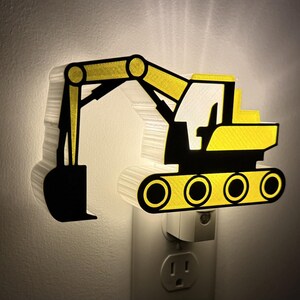 May include: A plug-in night light shaped like a yellow and black excavator construction vehicle. The light emits a soft glow from behind the silhouette, highlighting the detailed arm, bucket, cab, and track components of the heavy machinery.