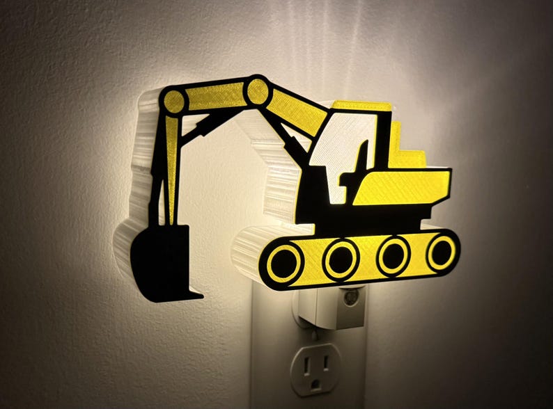 May include: A plug-in night light shaped like a yellow and black excavator construction vehicle. The light emits a soft glow from behind the silhouette, highlighting the detailed arm, bucket, cab, and track components of the heavy machinery.