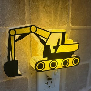 May include: A yellow and black excavator-shaped night light plugged into a wall socket. The construction vehicle design features a prominent bucket arm, a cab, and tracked wheels, emitting a warm, soft glow from its translucent yellow body.