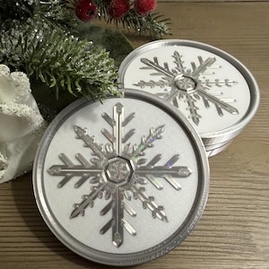 May include: Two round coasters with a silver snowflake design on a white background. The coasters have a silver rim and are stacked on a wooden surface. Decorative greenery and berries are in the background.