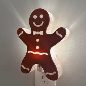 May include: A brown gingerbread man shaped night light plugged into a wall outlet. The figure features white icing details including eyes, a smile, a bow tie, two buttons, and wavy lines on the arms and legs. The light emits a warm glow.