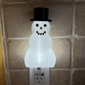 May include: A white snowman night light with a black top hat, black eyes, a carrot nose, and a smiling mouth. The night light is plugged into a wall outlet. The snowman is a festive decoration.