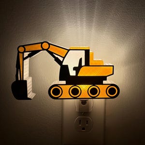 May include: A yellow and black excavator-shaped night light plugged into a wall socket. The construction vehicle design features a long articulated arm with a bucket, a cab, and tracked wheels, emitting a warm glow from behind the structure.