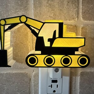 May include: A yellow and black excavator-shaped night light plugged into a wall socket. The construction vehicle design features a prominent bucket arm, a cab, and tracked wheels, emitting a soft glow from behind the structure to illuminate the area.