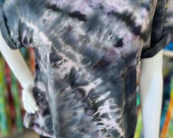 May include: A short-sleeved, tie-dye t-shirt in shades of gray, black, and purple. The shirt has a crew neck and rolled-up sleeves. The tie-dye pattern creates a marbled effect across the fabric. The shirt is displayed on a mannequin.