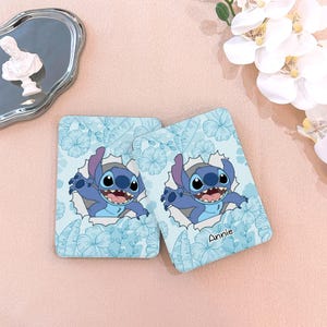 May include: Two light blue coasters featuring a cartoon character, Stitch, bursting through a torn paper effect. The coasters have a floral pattern and the name "Annie" is printed on one. The background is a light pink.