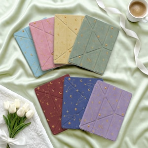 Può includere: Una collezione di sette custodie rettangolari per tablet in vari colori, tra cui blu, rosa, giallo, verde salvia, rosso scuro, blu navy e lavanda. Ogni custodia presenta un design pieghevole geometrico e motivi celesti dorati di stelle e lune.