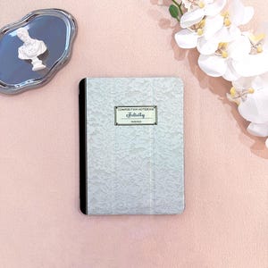 Puede incluir: Un cuaderno de composición azul claro con una cubierta de encaje. El cuaderno tiene un lomo negro y una etiqueta que dice "Composition Notebook Felicity". El cuaderno está sobre una superficie rosa.