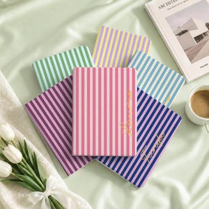 May include: A collection of five rectangular notebooks featuring vertical stripes in various colours including pink, purple, green, yellow, and blue. Two of the notebooks display gold cursive text on the front covers. The notebooks are arranged in a fan shape.