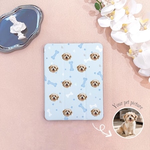 Puede incluir: Una funda de tableta azul claro con un patrón de caras de perro y formas de hueso azules. La funda tiene una foto de un perro en un círculo con el texto "Your pet picture". La funda está sobre una superficie rosa.