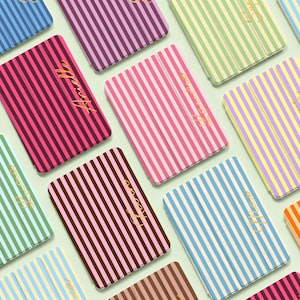 Op de afbeelding: Een collectie rechthoekige tablethoezen met verticale strepen in diverse kleurencombinaties zoals roze, blauw, groen, paars en bruin. Elke hoes is voorzien van een in goudkleurig cursief schrift gedrukte naam.