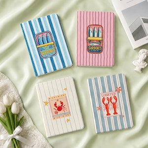 May include: Four rectangular notebooks with striped covers. One is blue-striped with a sardine tin graphic, one is pink-striped with a sardine tin graphic, one is cream-striped with a crab graphic, and one is blue-striped with a lobster graphic.