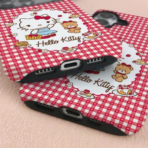 May include: Two black phone cases with a red and white gingham pattern. Each case features a white oval with Hello Kitty and a teddy bear illustration. The cases have cutouts for the camera and charging port.