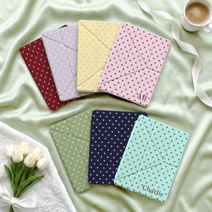 May include: A collection of seven rectangular tablet cases featuring a polka dot pattern in various colors including maroon, lavender, yellow, pink, sage green, navy blue, and mint green. Two cases feature personalized text, AB and Charlie.