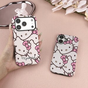 May include: Two white phone cases with a repeating pattern of Hello Kitty faces and pink bows. One case is held in a hand, displaying the name "Gabriel" in script. The cases have black camera cutouts and are on a pink surface.