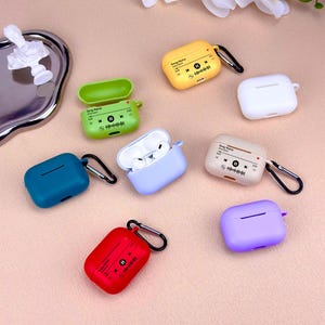 May include: A selection of brightly coloured AirPod cases in shades of yellow, green, blue, red, white, and purple. Some cases feature a music player interface design. Each case has a black carabiner clip.