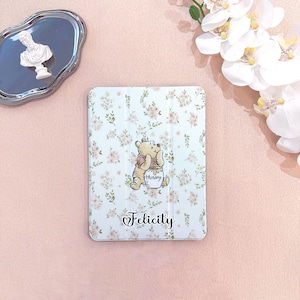 May include: A white tablet case with a floral pattern and a cartoon image of Winnie the Pooh holding a honey pot. The name "Felicity" is written in cursive below the image. The case is on a pink surface.
