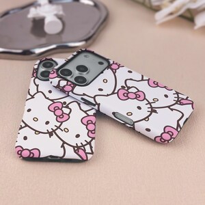 May include: Two white phone cases with a repeating pattern of Hello Kitty faces and pink bows. The cases have a black camera lens cutout and a black border. The design features black outlines and pink accents.