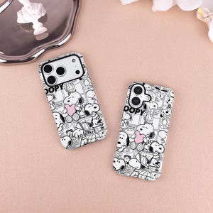 Könnte beinhalten: Zwei weiße Handyhüllen mit einem schwarz-weißen Comic-Design mit Snoopy, der ein rosa Herz hält. Die Hüllen haben eine glänzende Oberfläche und einen schwarzen Kameraausschnitt. Das Wort "DOPY" ist oben auf jeder Hülle aufgedruckt.