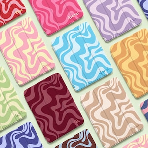 Puede incluir: Una colección de fundas rectangulares para tabletas con un patrón ondulado y jaspeado en varias combinaciones de colores, incluyendo rojo, rosa, azul, morado, amarillo y marrón. Varias fundas están personalizadas con nombres como Elise, Emma y Lisa.