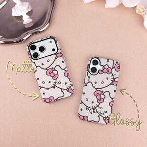 May include: Two phone cases with a Hello Kitty design. One case has a matte finish, and the other has a glossy finish. Both cases feature a white background with pink Hello Kitty faces and bows. One case has the name "Gabriel" written on it.