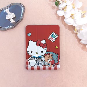 Puede incluir: Funda roja para tableta con Hello Kitty leyendo un libro, con un osito de peluche y la palabra "Felicity". La funda tiene un patrón de cuadros rojos y blancos en la parte inferior, con detalles de corazón azules y amarillos. También aparece un libro de "Cuentos de hadas".