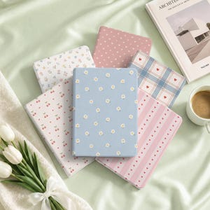Puede incluir: Una colección de seis cuadernos rectangulares con varios patrones, incluyendo margaritas blancas sobre azul claro, cerezas rojas sobre blanco, corazones rosas sobre rosa claro, cuadros azules con corazones rojos y estampados florales pequeños.