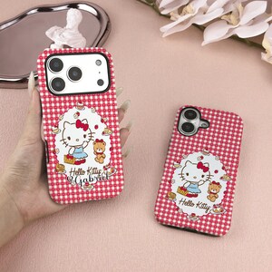 May include: Two red and white checkered phone cases. One case is held in a hand. Each case features a Hello Kitty design with a red bow, a teddy bear, and the text "Hello Kitty Gabriel" or "Hello Kitty".