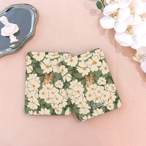 May include: Two square coasters with a floral design and a squirrel peeking out from the flowers. The coasters have a green background with white flowers and a squirrel in the center. The name "Annie" is printed on one of the coasters.