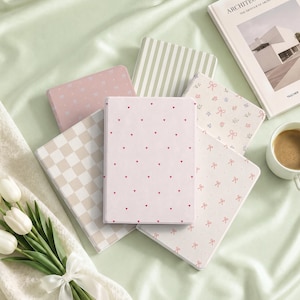 May include: A collection of five rectangular notebooks featuring various patterns including a light pink cover with small red hearts, a beige and white checkered design, a soft pink cover with white bows, a cream cover with green stripes, and a floral bow print.
