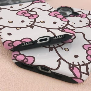 May include: Two white phone cases with a repeating pattern of Hello Kitty's face, featuring pink bows and brown outlines. The cases have black edges with speaker and charging port cutouts.