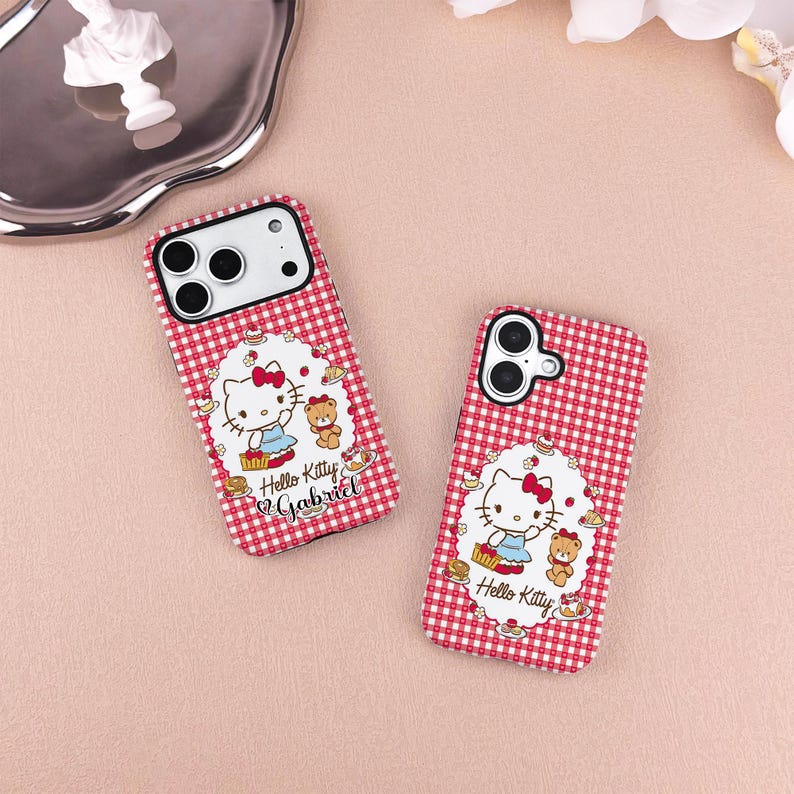 May include: Two red and white checkered phone cases featuring a Hello Kitty design. Each case has a white oval with Hello Kitty, a teddy bear, and the text "Hello Kitty Gabriel". The cases are on a pink surface.