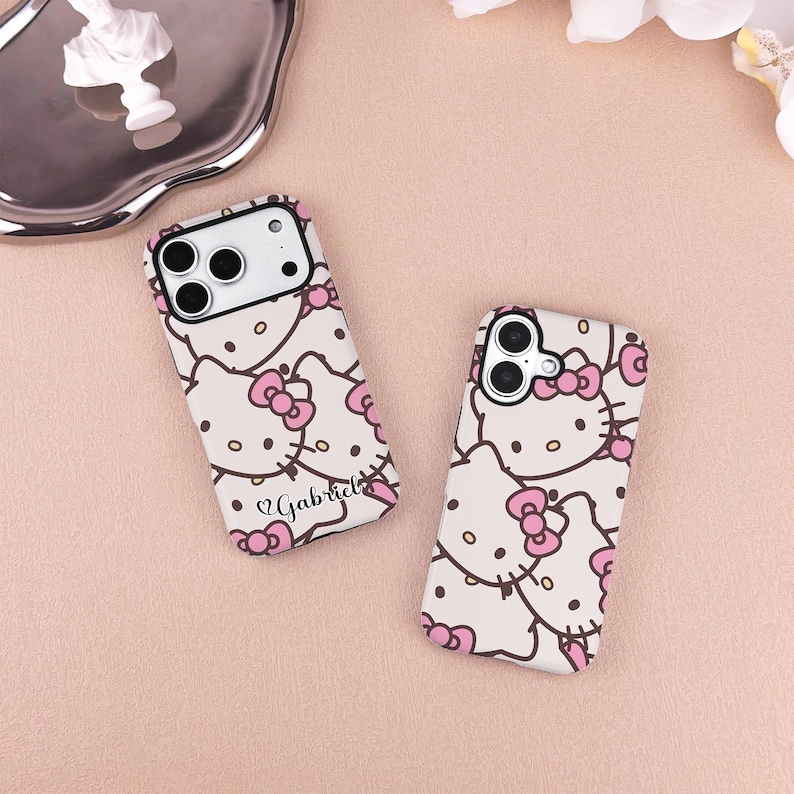 May include: Two phone cases with a repeating pattern of a cartoon cat with a pink bow. The cases are white with pink accents and the name "Gabriel" is printed on one of the cases.